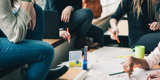 Brainstorming Vier Menschen sitzen an einem Tisch, die Gesichter sind nicht im Bild. Sie halten Stifte in der Hand und sehen nachdenkend aus. Eine Hand schreibt etwas auf das Plakat auf dem Tisch, welches sehr unstrukturiert gestaltet ist. Auf dem Tisch sind noch mehr Stifte und eine Tasse. Auf der Sitzbank erkennt man einen geöffneten Laptop und einen Rucksack im Hintergrund.